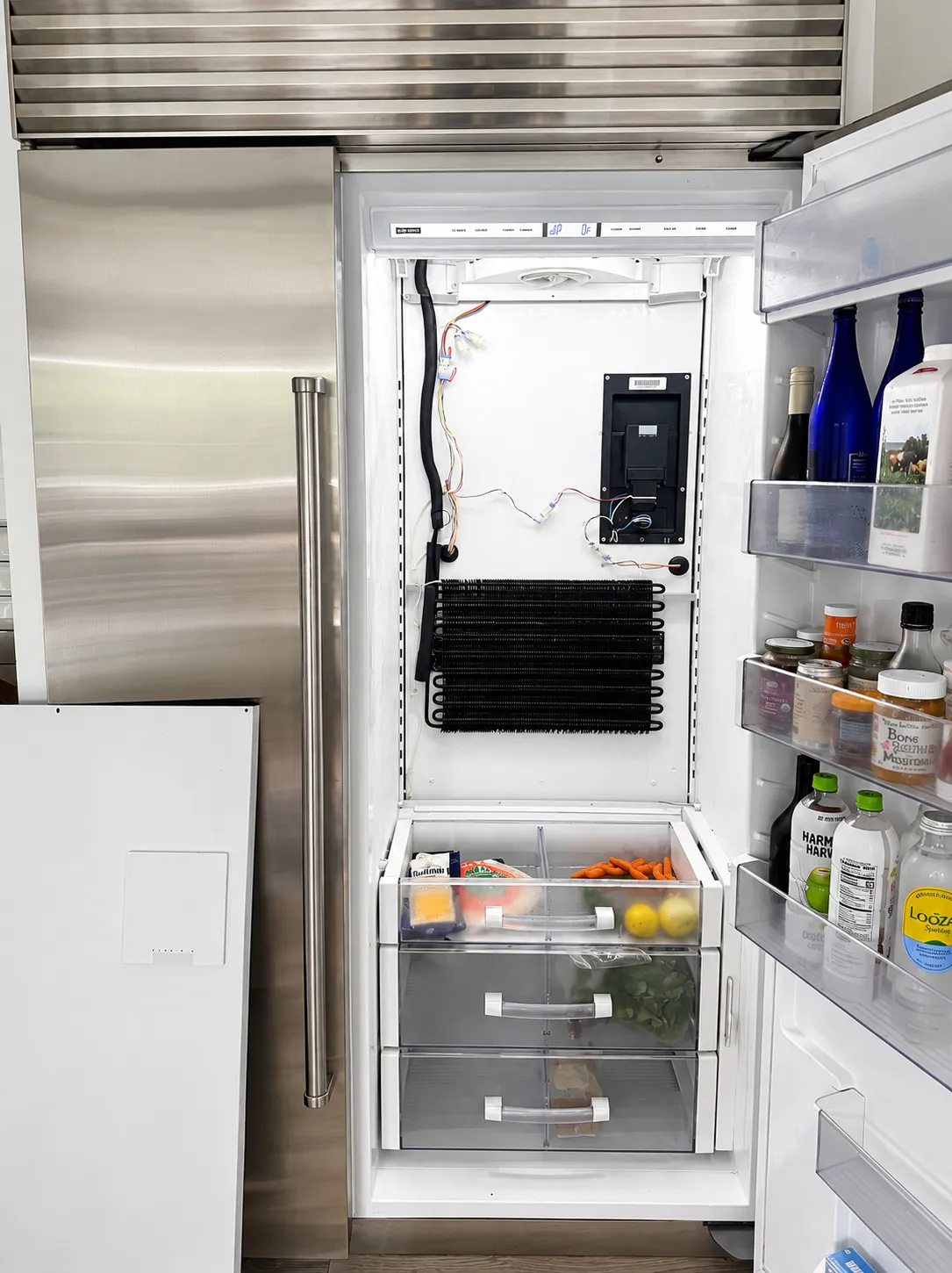 An open built in Sub-Zero refrigerator with the interior back panel removed, exposing the evaporator coil, control board, and wiring