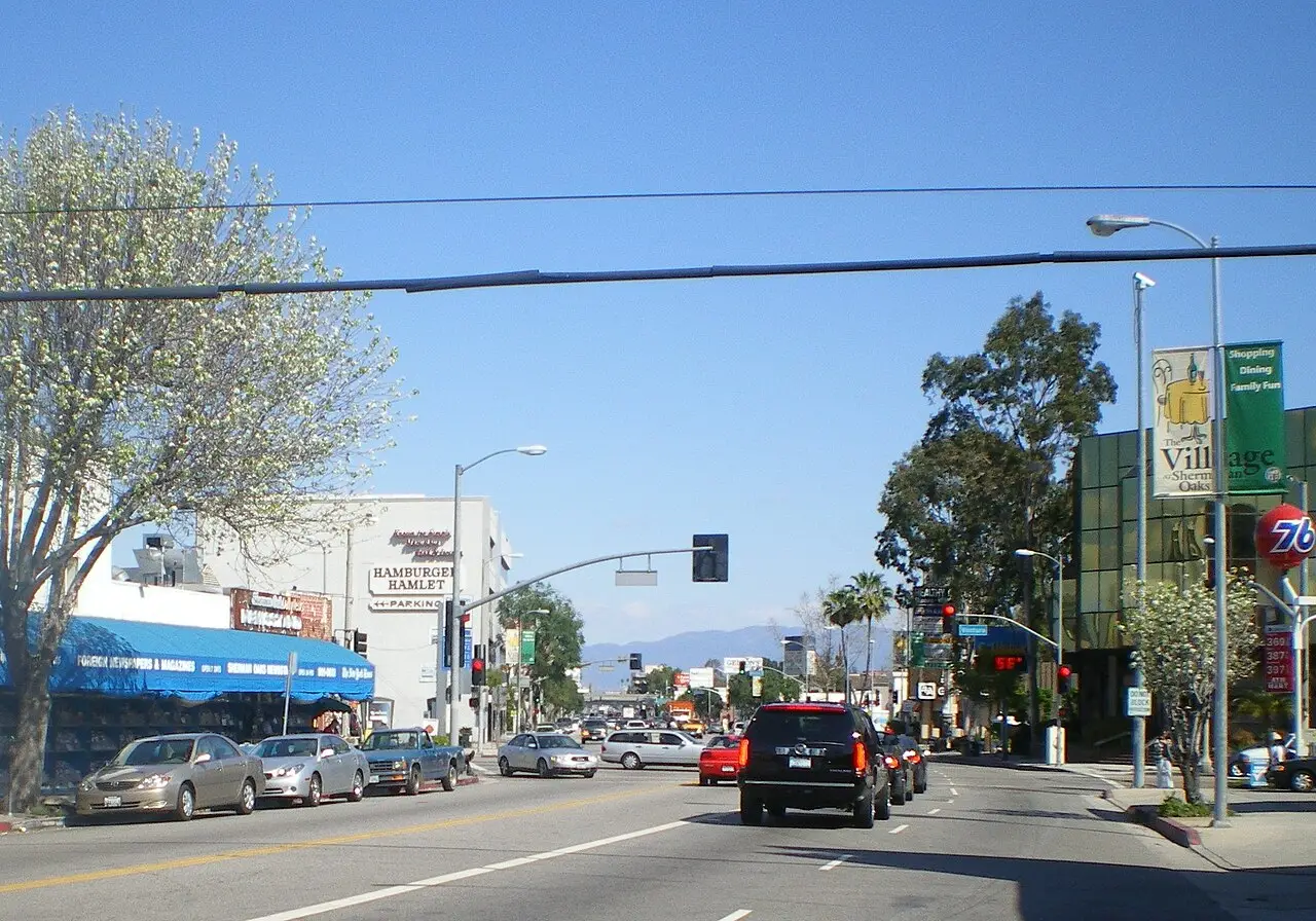 Storefronts and a newsstand along a busy Sherman Oaks boulevard, mountains in the distance