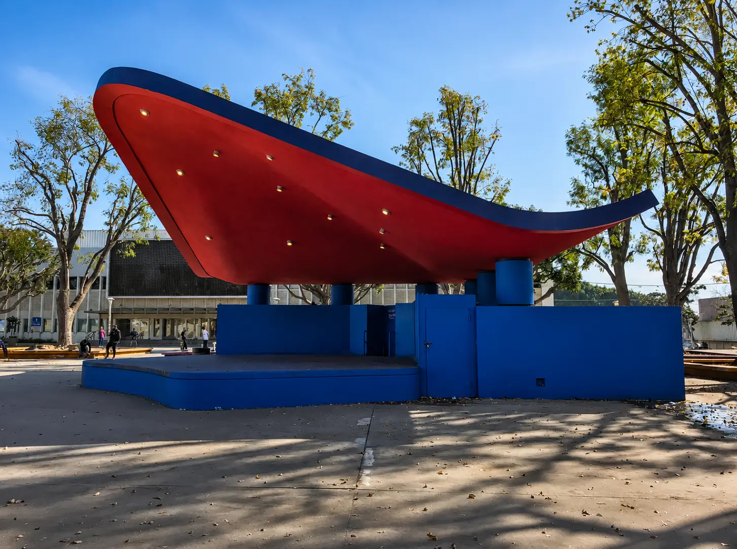 Red-and-blue modernist bandshell on a sunny tree-lined park plaza in West Los Angeles
